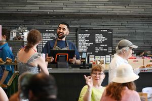 A person smiling behind a counter with customers milling around in the foreground.