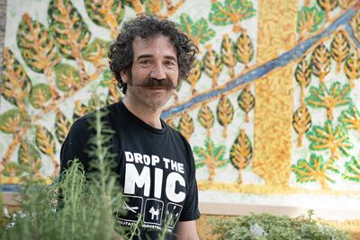 Michael Rakowits smiles in front of his artwork. He has dark curly hair and a moustache.