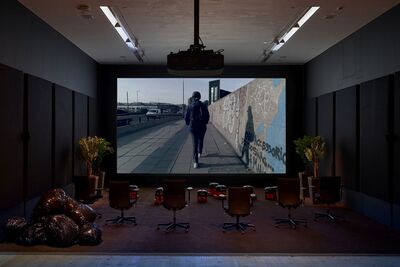 A gallery room is filled with office chairs, fun bin bags, and Henry Hoovers while a projected screen plays a film.