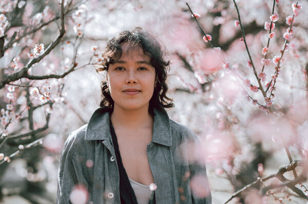 Woman with dark curly hair and a grey jacket stood amongst pink blossom trees.