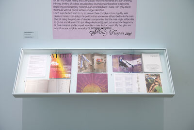A glass cabinet on a white wall displaying letters and magazines. There is a pink sign above with writing on it. 
