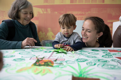 Boy adding to drawing of plants with a cut out shape. Grandma and mum are smiling next to him.