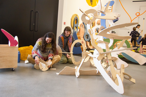 Two people sat on the floor playing with part of a wooden abstract sculpture.