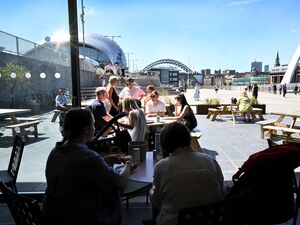 Group of people sat outside on a sunny day with views of the Gateshead quayside in the background.