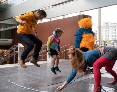 Visitors jumping over skipping rope with character wearing yellow and blue fringe costume