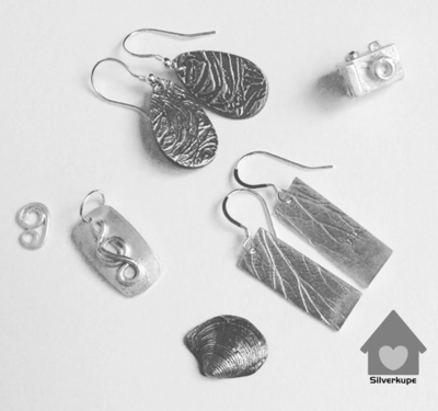 An array of silver jewellery including earrings, and small pedants in the shape of a camera, shell, and music note.