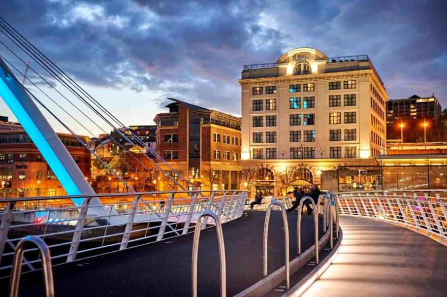 A grand looking hotel lit up in the evening behind a river and bridge.