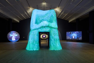 A large sculpture of a body with a doorway through the middle leading to an eye. There are vibrant blue projections on the sculpture. Either side of the sculpture there are screens displaying animations.