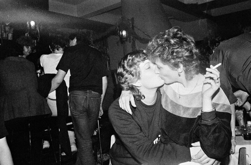 Black and white film still of two people kissing in a bar. One of them is holding a cigarette.