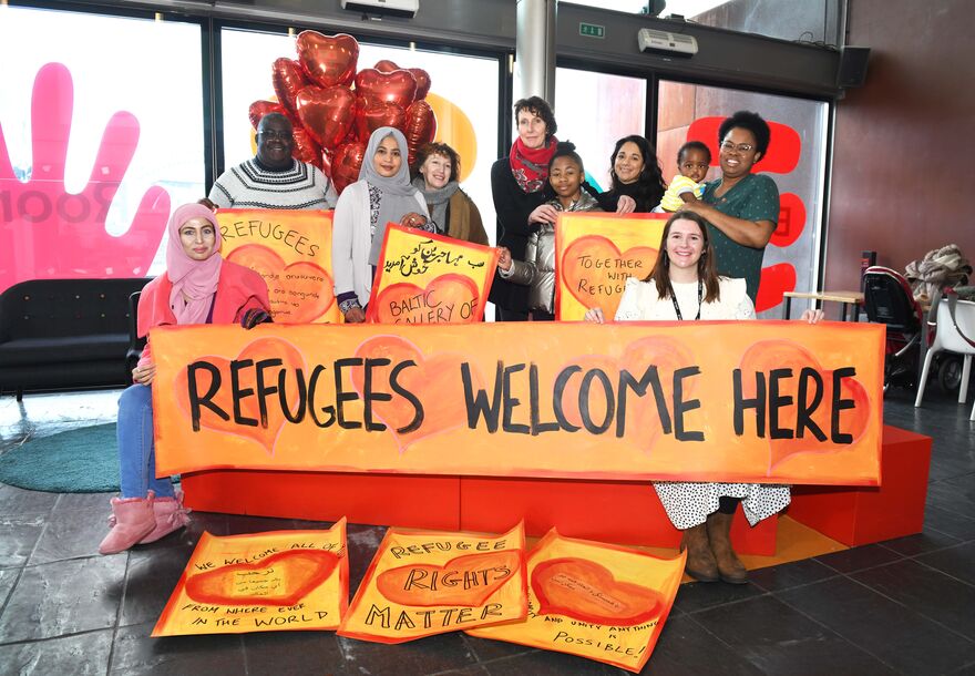 Group of people stood together smiling and holding up a big orange sign that reads 'refugees welcome here'