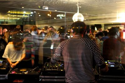 Back of a man wearing a stripey top and headphones stood at a DJ deck. In the background there's a crowd of people and a disco ball hanging over them.