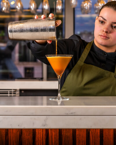 Woman wearing a dark green apron pouring cocktail mix into a glass.