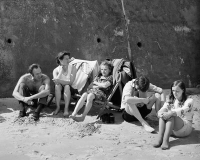 Black and white photo of five adults sat on the beach. Two of them are on wooden beach chairs.