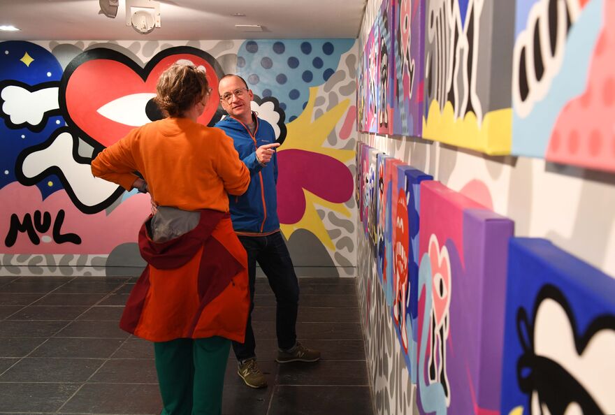 Two people standing in Mul's pop up shop at baltic in front of a mural, pointing at original paintings hung on the wall.