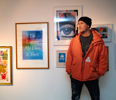 Man wearing an orange coat and black beanie stood against a wall covered in framed collages.