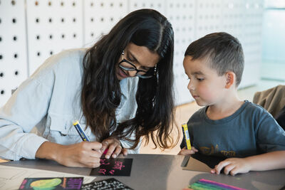 Woman with long black hair and glasses writing with felt tip next to a young boy who's watching.