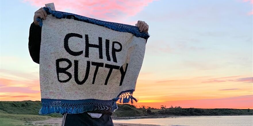 Person holding fabric sign on seafront saying 'Chip Butty'
