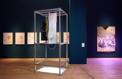 A metal scaffold sculpture with screens and wires inside it. In the background is a large dark blue wall with artwork displayed on it.