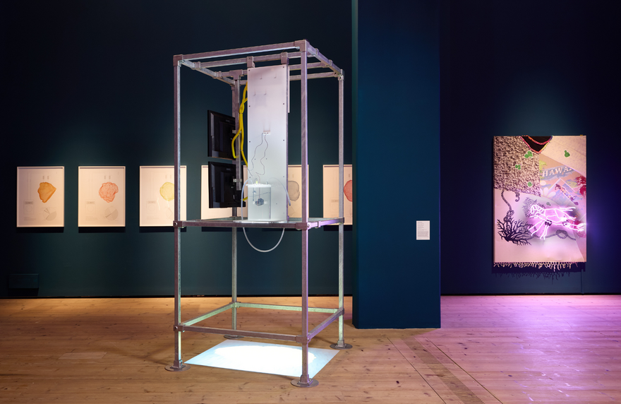 A metal scaffold sculpture with screens and wires inside it. In the background is a large dark blue wall with artwork displayed on it.