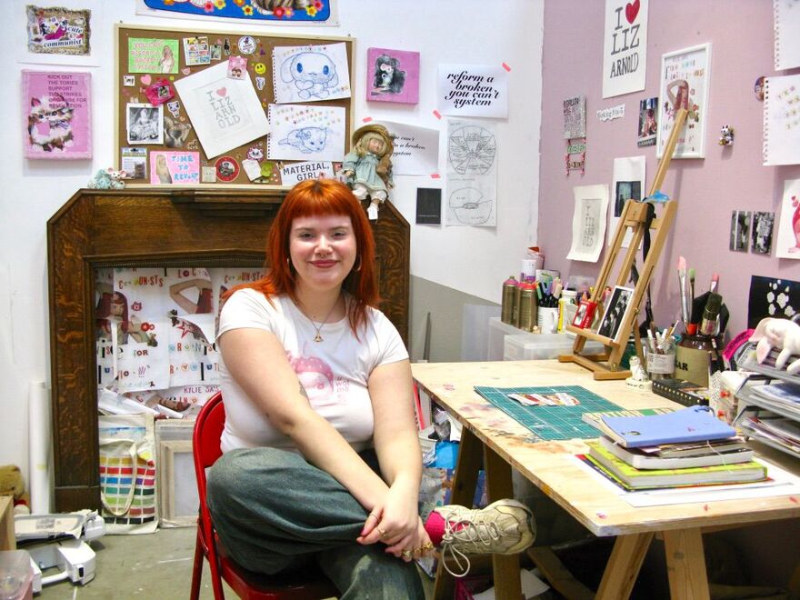 Elleanna Chapman sat in her studio at a table with her legs crossed wearing a white graphic tshirt and grey trousers and trainers and she has straight red/orange hair with a fringe. She is surrounded by her art which is colourful, mainly pink and white with cute images of cats and Sanrio characters.