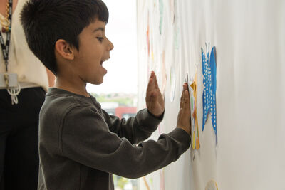 A young boy smiling and pressing cut out artwork onto a wall.