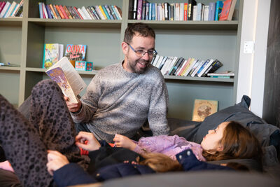 Girl lying on beanbag while dad smiles and reads to her. Behind them is a green bookcase full of books.