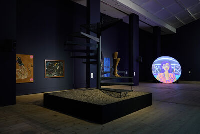 Metal staircase with a sculpture of legs descending on it. There are paintings on displayed on a navy wall behind this, as well as a circlar screen showing an animation.