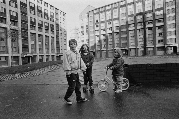 Three kids stood together with a bike in front of a block of apartments.