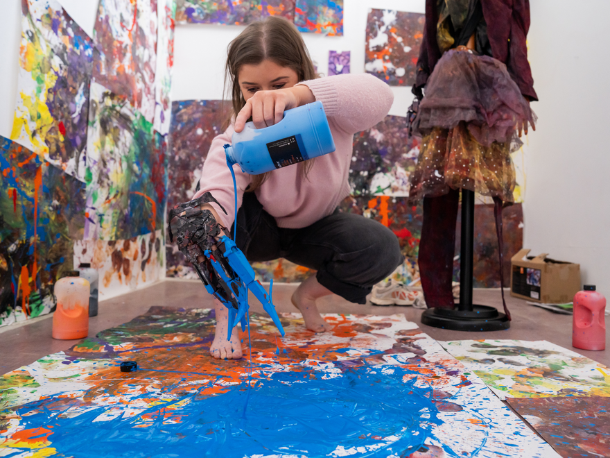 Person pouring bright blue paint onto colour-splattered paper on the floor.