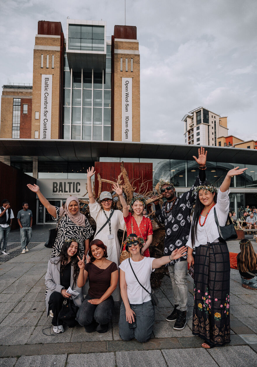 Group of people smiling and waving stood outside Baltic and wearing flower crowns.