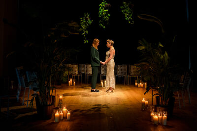 Two women stood holding hands in a room surrounded by green plants and candles.