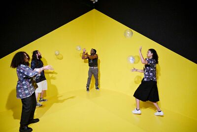 Group of four young people playing with large glittery floating bubbles against a black and yellow wall.