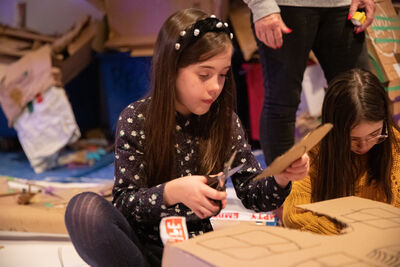 Girl with long brown hair and spotty headband sat cutting out a cardboard box.