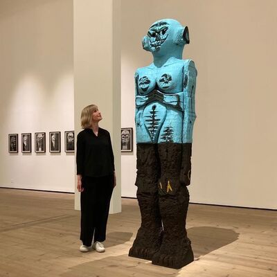 Woman wearing all black and white trainers, with short blonde hair, stood next to a big blue sculpture