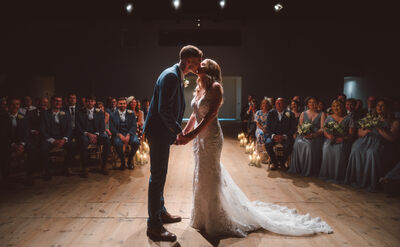 Two people in wedding attire kissing infront of a seated audience.