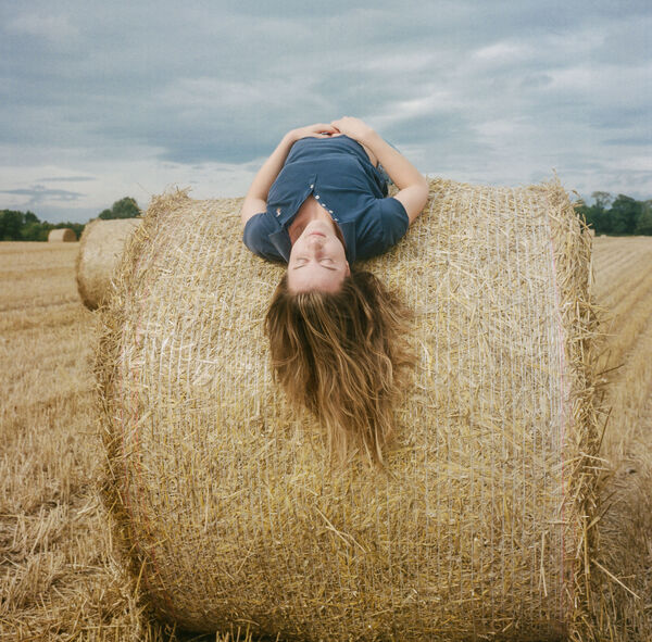 Person with long brown hair wearing a blue top lying on her back on top of a hay bale