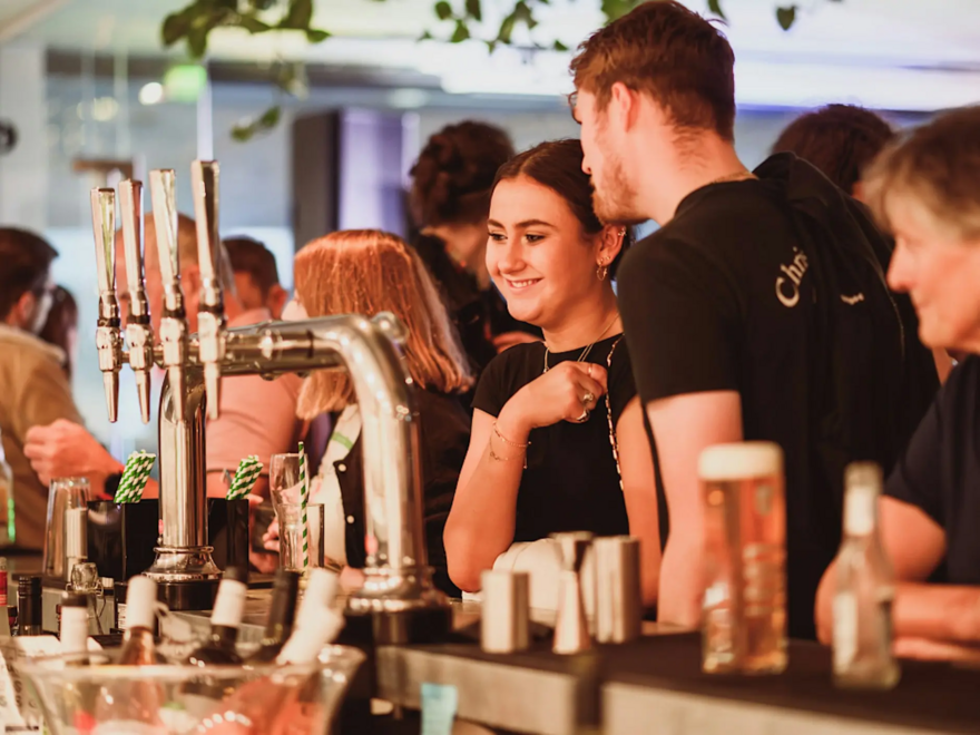 Group of people smiling and stood at a bar with a pint of beer in shot.