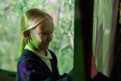 girl is in an exhibition with a green image projected onto the wall and her face, she is touching the walls of the exhibition with her hand and smiling