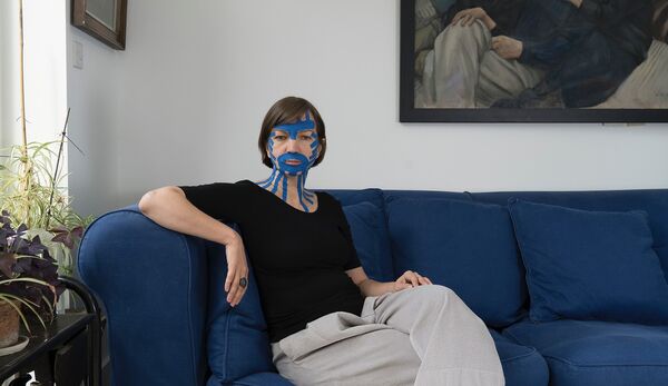 Woman with short brown hair wearing black T-shirt and grey trousers sat on deep blue sofa. She has blue tape over her face.
