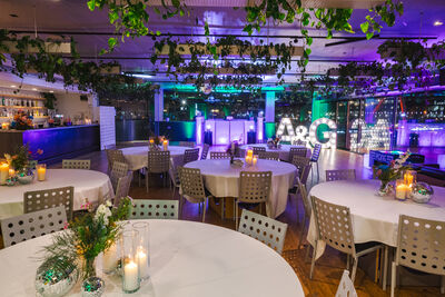 Room with round tables covered in white tablecloth, with purple lighting and green plants hanging overhead.