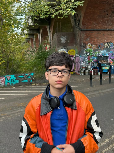 A young person with short brown hair, glasses and a vibrant orange jacket with headphones around their neck.