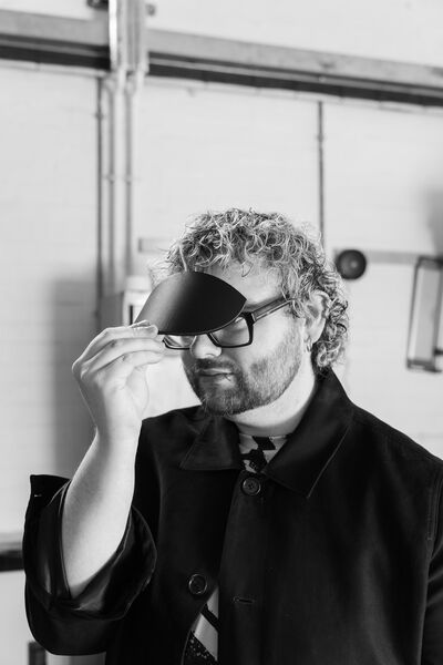 Black and white photograph of a man with light curly hair, glasses, a hoop earring and wearing a dark casual jacket.