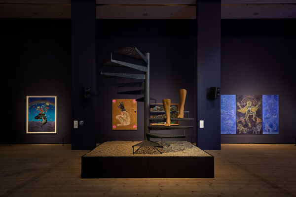 Metal winding staircase with sculpture installation of legs descending staircase. Behind is a selection of vibrant paintings displayed on a navy blue wall.
