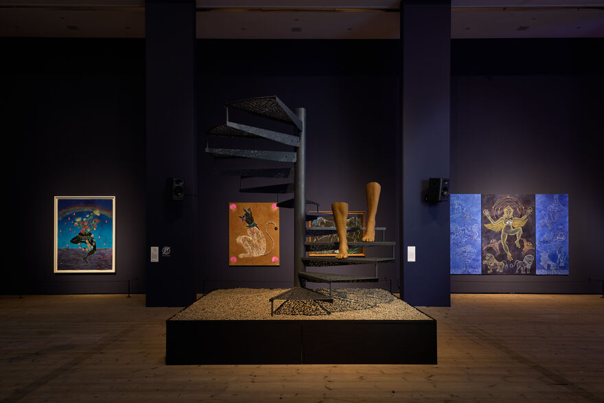 Metal winding staircase with sculpture installation of legs descending staircase. Behind is a selection of vibrant paintings displayed on a navy blue wall.