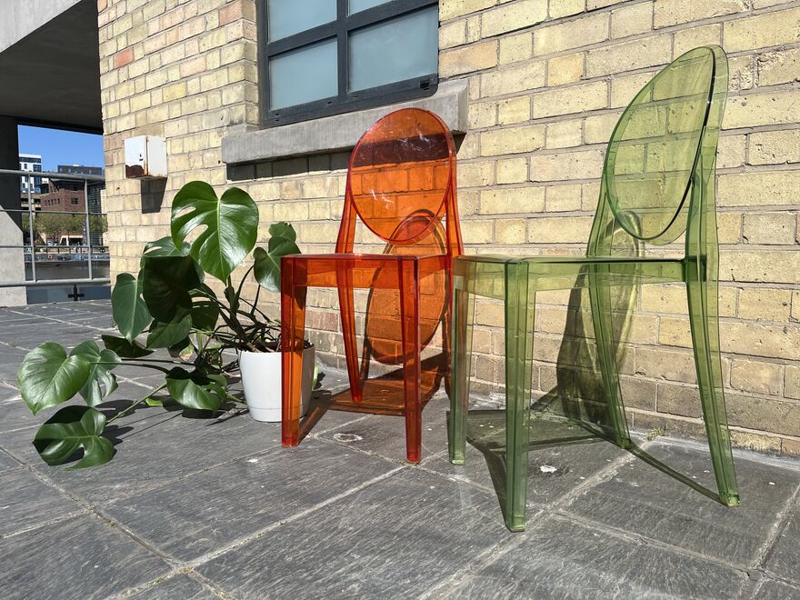 Two acrylic chairs, one orange and one green with a large monstera plant next to them, and against a brick wall.