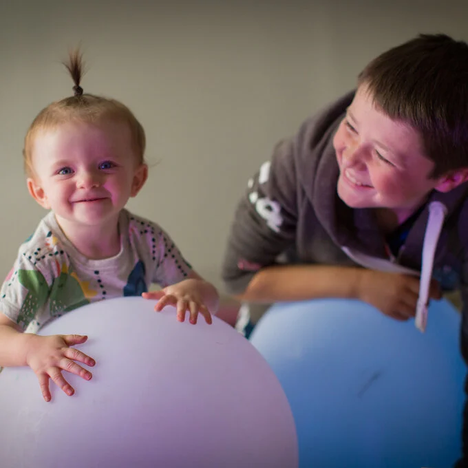 A smiling young child and older boy holding and lying on large and soft colourful balls. 