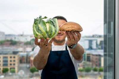 Person wearing a black apron and white t-shirt holding a cauliflower and a large mushroom infront of a city-view window,