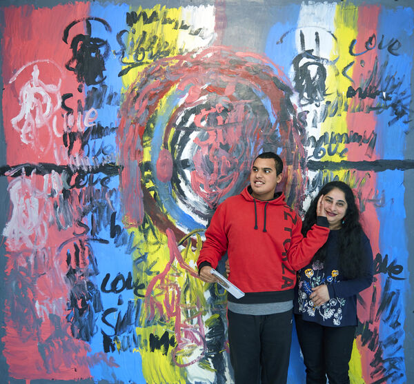 Two people stood in front of a colourful mural in red, yellow and blue.