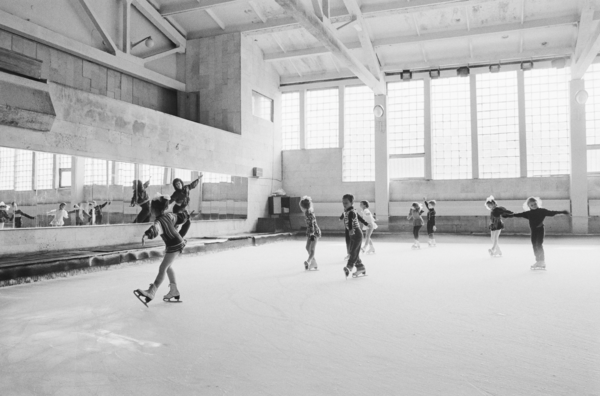 Black and white photo of a large industrial room with a woman at the front teaching a group of kids how to ice-skate.