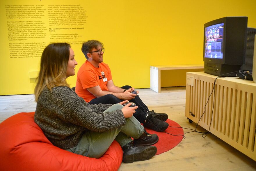 Two people sat on red beanbags, holding retro consoles and playing a game. There is a bright yellow wall in the background